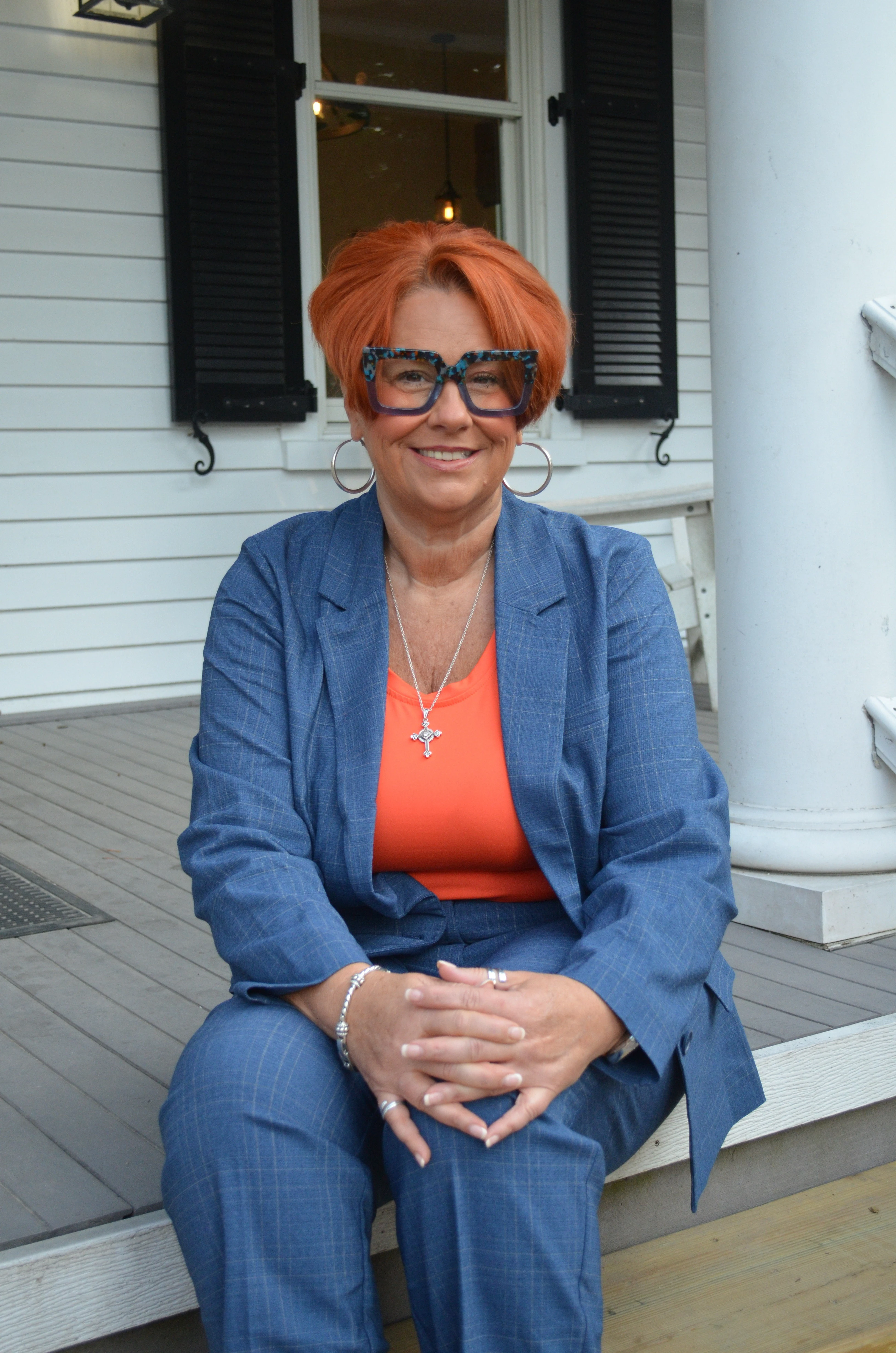 Corrina Carter, copper-orange hair and tortoise-shell glasses, sitting on the wooden steps of a white southern porch, hands clasped, warm natural light